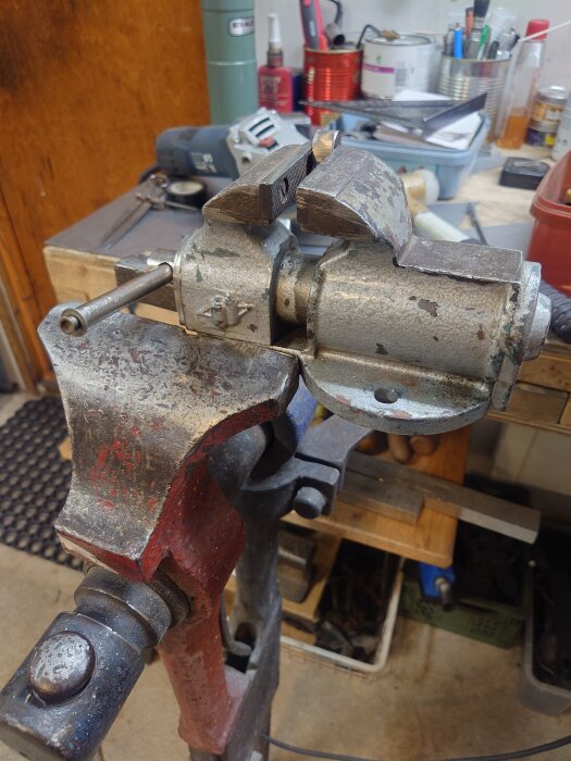 Bänkmonterat skruvstäd i verkstadsmiljö med verktyg och förvaringslådor i bakgrunden.