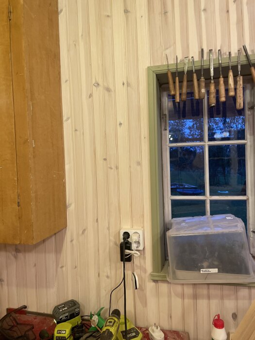 Vittlaserat trävägg med kabinetter och verktyg, fönster med grönram. Arbetshörna med verktyg och eluttag.