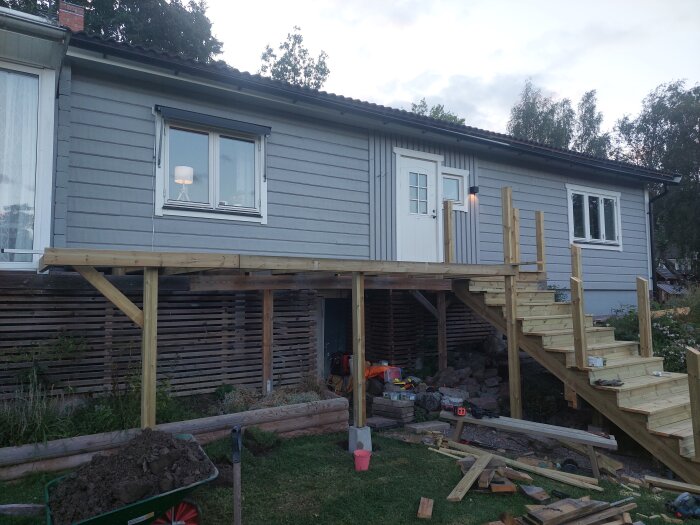 Byggprojekt med en nästan färdigställd altan eller balkong, nybyggd trappa och arbetande verktyg omkring; huset är grått med vit dörr.