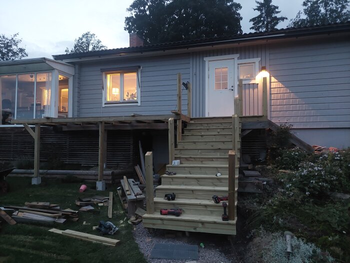Nybyggd trappa i trä leder upp till en altan eller balkong på ett hus med vita dörrar och fönster. Byggmaterial och verktyg syns i förgrunden.