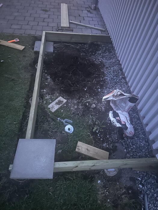 Grundkonstruktion för redskapsbod med träreglar och betongplattor på grus och jord, omges av en stenlagd yta och en byggnadsvägg.