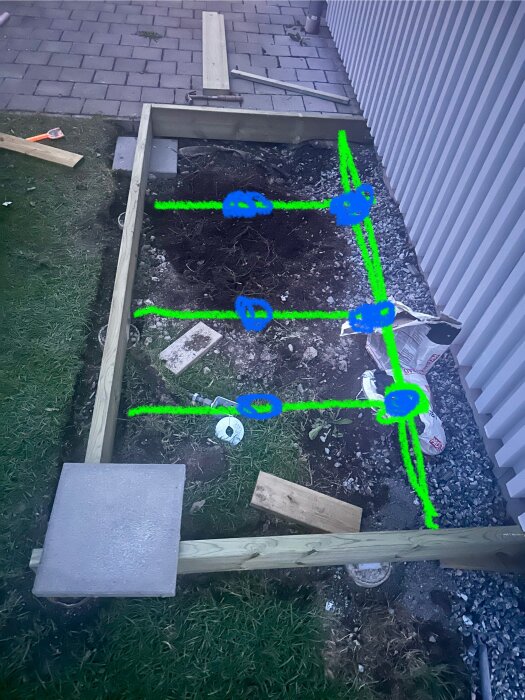 Byggram med korta reglar och en plint, markerad med blått och grönt, på gräs och stenplattor vid husvägg. Verktyg och träbitar synliga runtom.