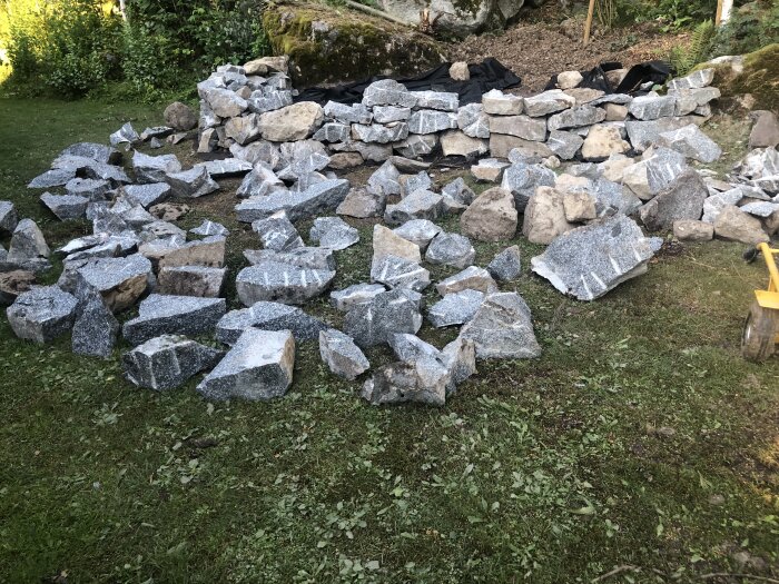 Stora stenar krossade i mindre block ligger på marken, omgivna av grönska. En mur av staplade stenar i bakgrunden.