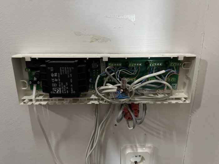 Elektrisk kopplingsbox för golvvärmesystem, med synliga kablar och kretskort mot en vit vägg.