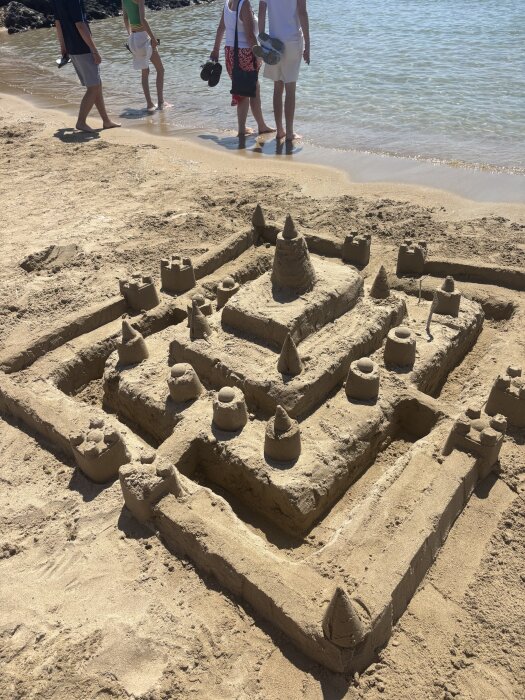 Sandslott på stranden med flera torn och murar, havet i bakgrunden och fyra personer står vid vattenbrynet.