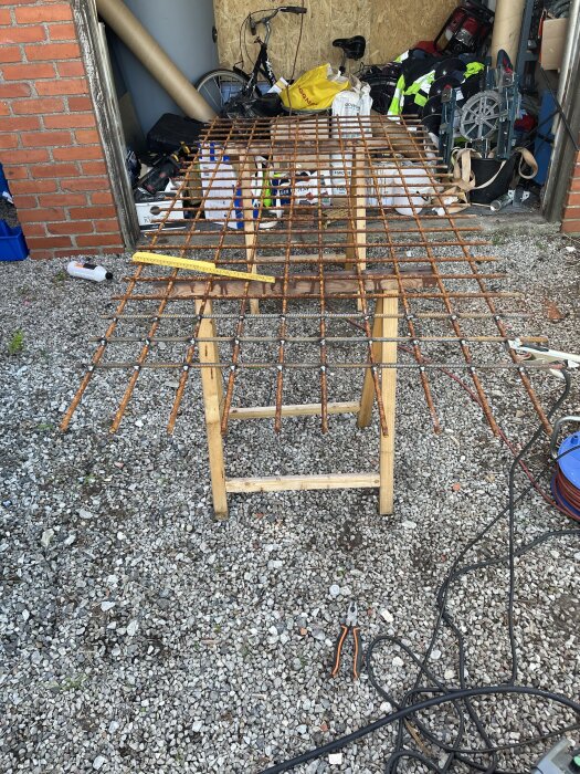 Ameringsnät ligger på träbockar med svets utrustning och verktyg runt, i en verkstadsmiljö med byggmaterial och cykel i bakgrunden.