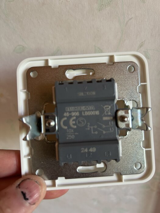 Närbild på en grå kronbrytare från Biltema, sedd framifrån mot en vit vägg. En hand håller i brytaren.