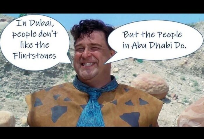 En man i stenåldersklädsel med pratbubblor: "In Dubai, people don't like the Flintstones. But the people in Abu Dhabi Do." I stenig utomhusmiljö.