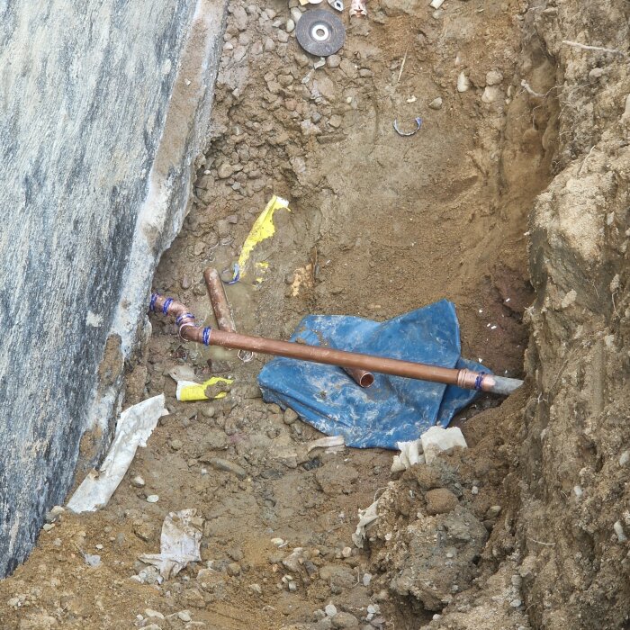 Grävd grop med exponerat kopparrör fullt av skarvar, omgiven av jord och plastskräp.