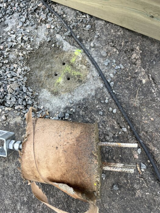 Borttagen betongplint med armeringsjärn, hål i marken för plintens återplacering, omgiven av grus och en träplanka.