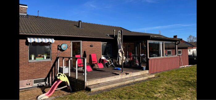 Rött tegelhus med soldäck och leksaksslide, inglasad veranda ses till höger, klarblå himmel. Lågt sluttande tak som diskuterats för renovering.