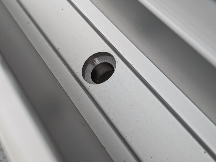 Närbild av aluminiumskena med stort, koniskt borrhål för montering på balkong, relaterad till glasräcke och skruvval.
