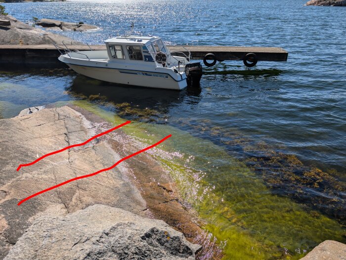 Båt vid brygga och klippa med markerade linjer för planerad båtslip, vatten med tång och solljus som reflekteras.