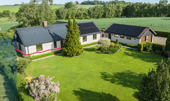 Ett hus med svart tak och tegelväggar, omgiven av grön trädgård och träd. En röd linje markerar en del av husets fasad.