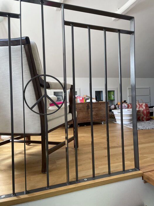 En metallräcke med en cirkeldetalj, står vid en övervåning med trägolv. I bakgrunden syns en stol och annan inredning.