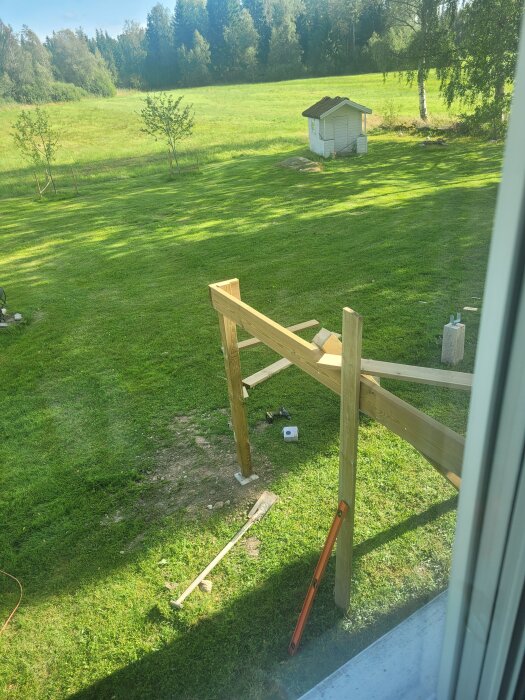 Påbörjad altanbyggnation i trädgård med träramar och verktyg i gräset, omgiven av grönska och med förråd i bakgrunden.