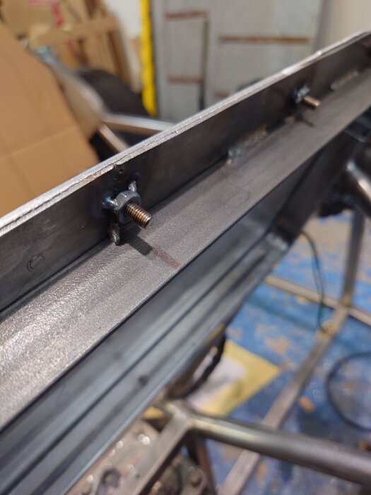 Svetsade muttrar på en metallbalk, verkstad i bakgrunden.