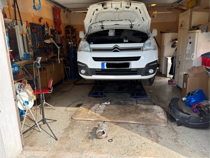Vit bil på billyft i garage, motorhuven öppen. Verktyg syns på väggarna. Markskydd och delar på golvet, garage fyllt med utrustning.