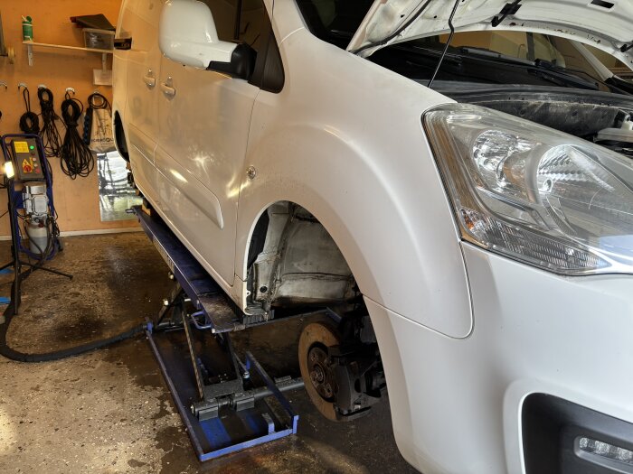 Vit bil på billyft med öppen huv, avlägsnade innerskärmar och arbeten med remmar och vattenpump i garage.