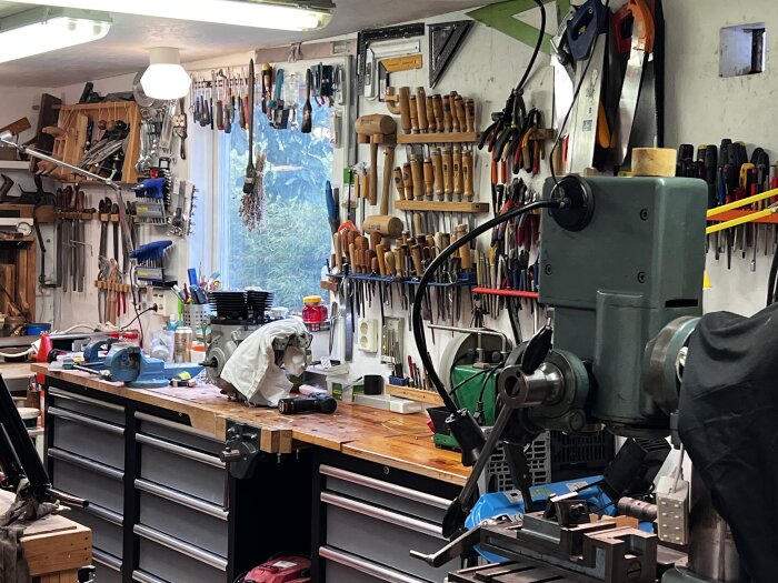 Verkstad med arbetsbänk full av verktyg, mejslar och tänger organiserade på väggarna, samt en maskin och motor på bänken.