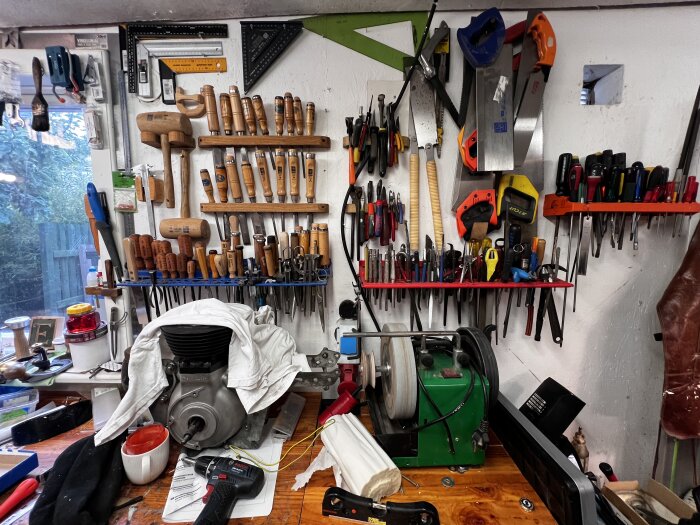 Verkstad med organiserade verktygshyllor fyllda med mejslar och sågar ovanför bänk. Framför bänk ligger en motor delvis täckt med en trasa.