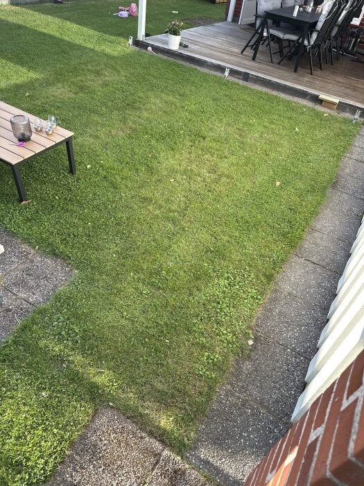 Gräsmatta med stenplattor runtom, intill en veranda med trämöbler. Plan för att bygga ut altanen med reglar och plankor diskuteras.