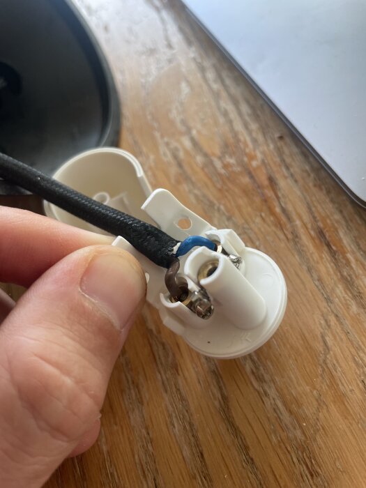 En hand håller i ett vitt DCL-lampdon med en svart sladd ansluten, placerat på ett träbord.