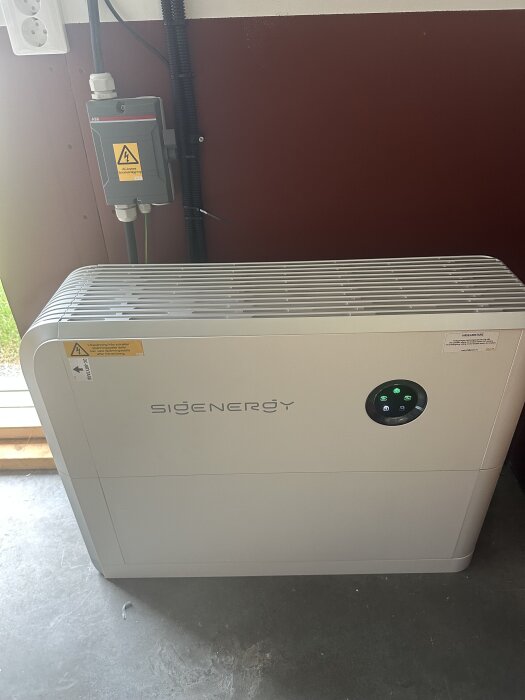 Vitt, rektangulärt Sigenergy-batteri på ett golv med en elbox på väggen bakom.