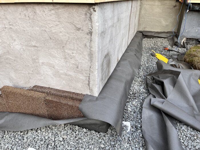 Bild av en husgrund med synliga dräneringsskivor och makadam täckt med fiberduk, omgiven av byggmaterial och verktyg.