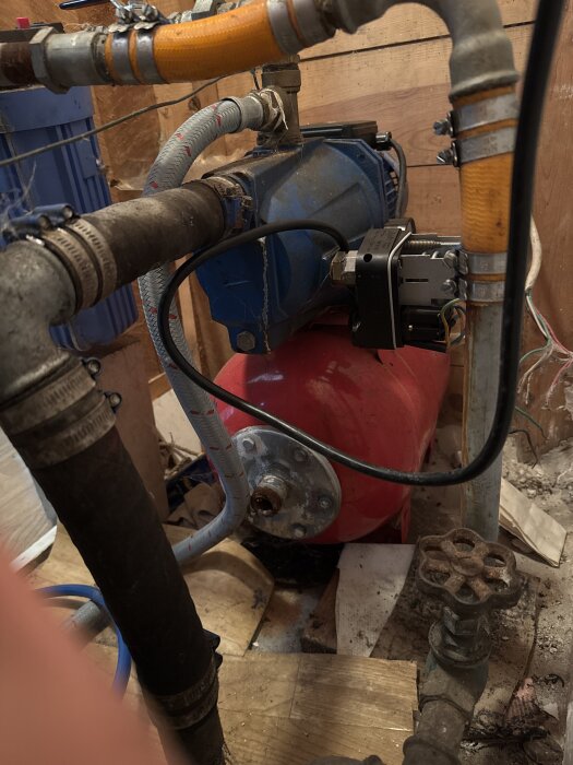 Pumphus med blå pump och röd trycktank omgiven av olika rör och ventiler i ett trångt utrymme med träväggar.