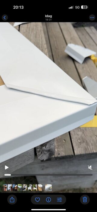 Vit hörnprofil i metall placerad på ett träbord utomhus, med ytterligare materialdelar i bakgrunden.