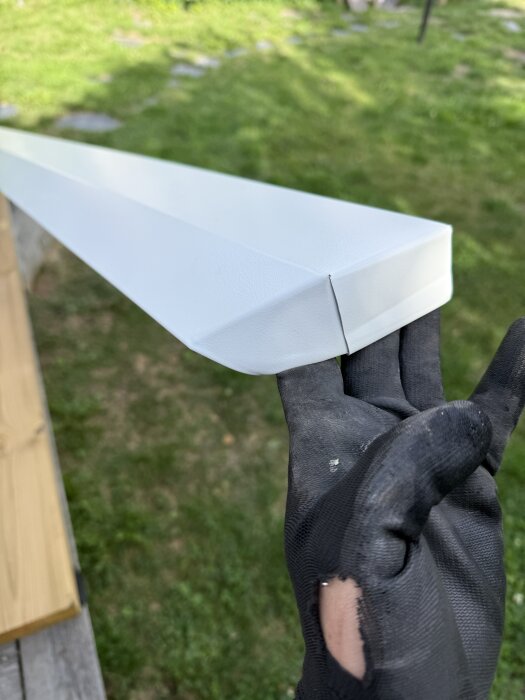 Hand håller en vit metallplåt med avslut och vinkel, troligen från ett byggprojekt, mot en grönskande bakgrund.