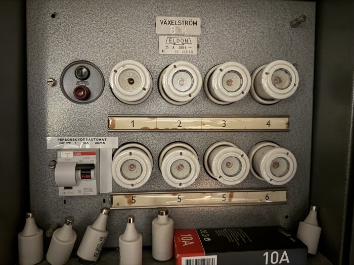 Elcentral med sex porslinssäkringar och en jordfelsbrytare, märkt "Växelström" och "Eldon", mot en grå bakgrund.