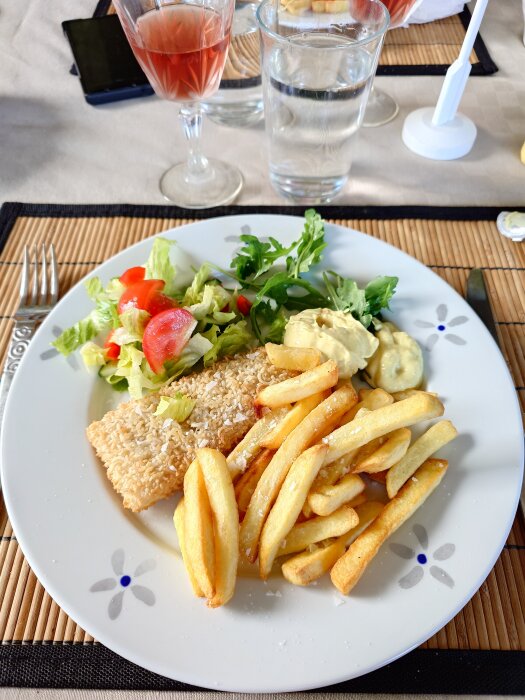 Tallrik med panerad fisk, pommes frites, sallad och sås. Ett glas rosévin och ett glas vatten syns i bakgrunden.