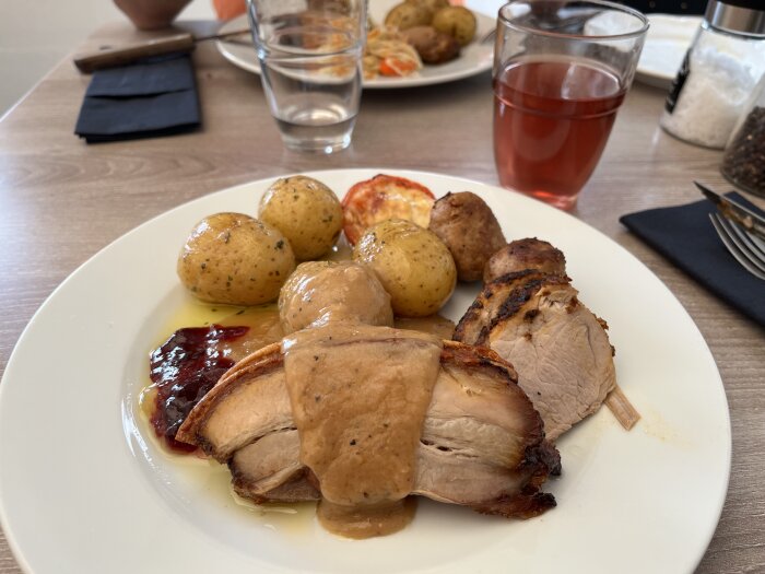 Tallrik med kött, sås, potatis och gelé, serverad med ett glas röd dryck på ett bord med bestick och servett.