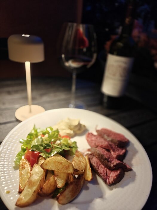 Tallrik med köttskivor, ugnsrostad potatis, sallad och sås. Vinflaska och glas i bakgrunden på ett dukat bord med lampa.