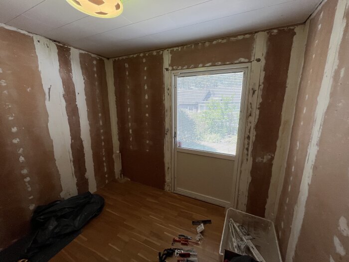 Ett rum under renovering med en altandörr och blottade väggar. Verktyg och plasttäcken syns på golvet, ljus strömmar in genom fönstret.