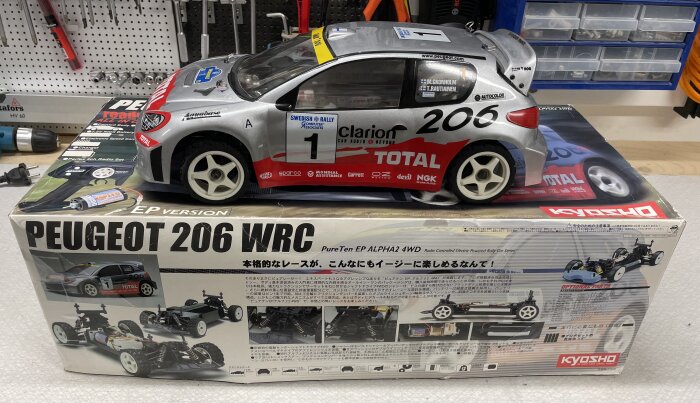 RC-bil Peugeot 206 WRC-modell från Kyosho på en verktygsbänk, vid sidan av dess originalförpackning med specifikationer och bilder.