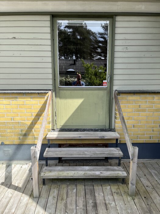 Grön altandörr med glasruta, trätrappa framför och spegelbild av en person som tar foto, sett på gul tegelfasad.