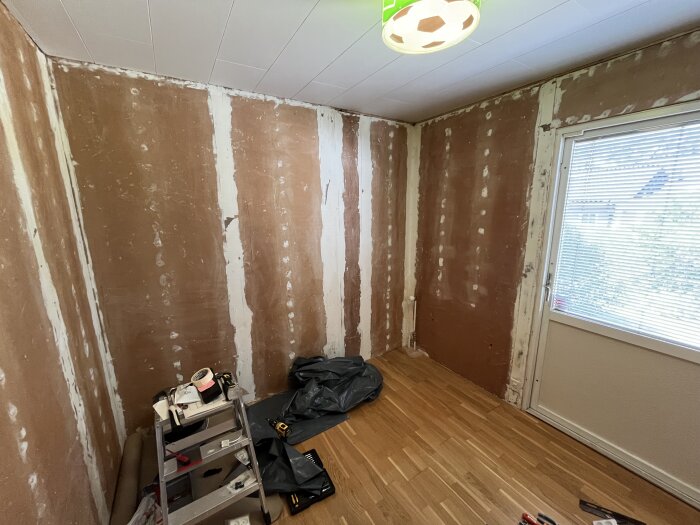 Ett rum under renovering med omålade väggar och trägolv. En stege med verktyg står mot en vägg, och en altandörr med persienner syns till höger.