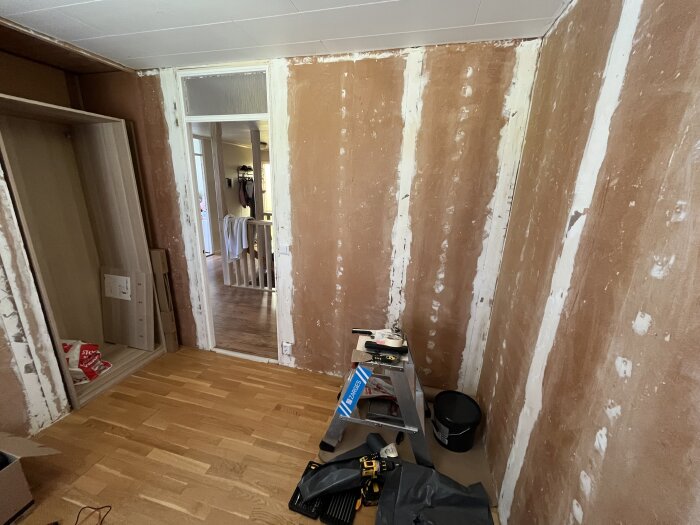 Ett rum under renovering med trägolv, en stege, verktyg och kartonger. Dörröppning till angränsande rum synlig.