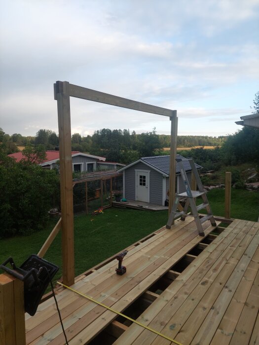 Pergolabygge med upprättstående stolpar på ett trädäck, stege och verktyg synliga, trädgård och friggebod i bakgrunden.