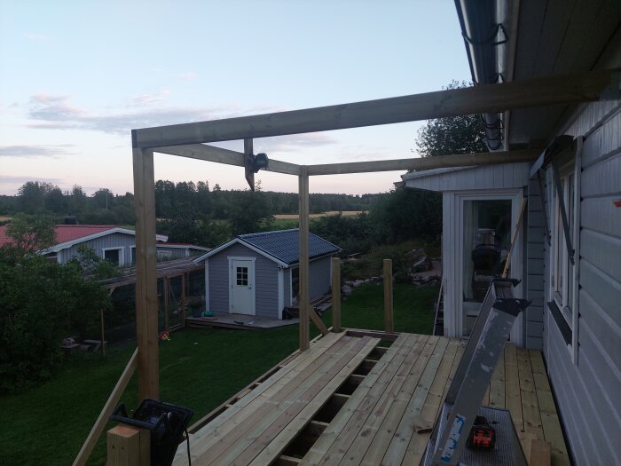 Pergolastomme byggd intill hus med stolpar och tvärgående reglar, provmonterad med stående trallvirke. Bostad och trädgård i bakgrunden.
