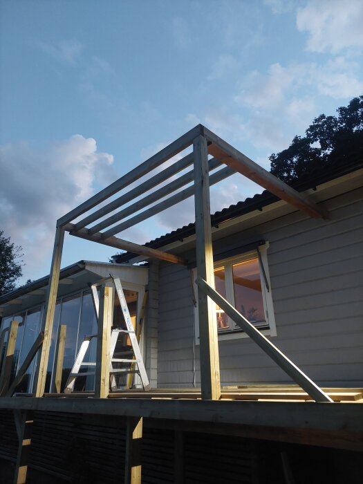 Pergolastomme under uppförande vid ett hus, med vertikala och horisontella träbalkar. En stege lutar mot konstruktionen.