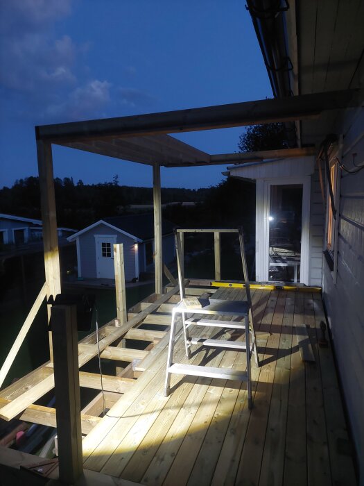 Bygget av en pergola med monterade stolpar på en altan, kvällstid med bygglampa och stege.