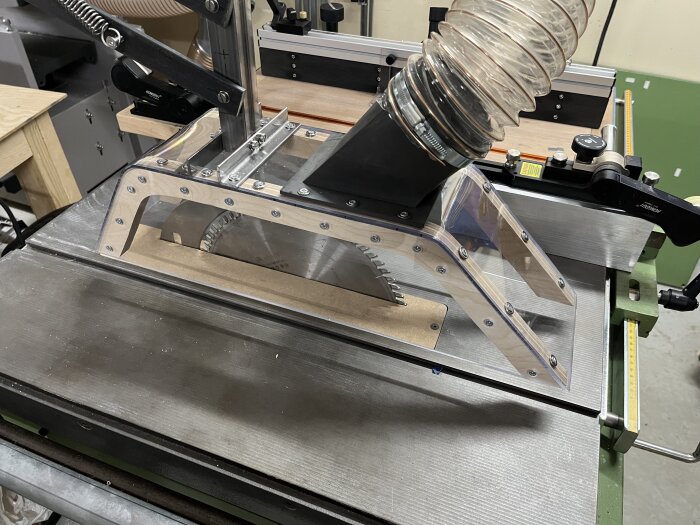 Specialbyggt klingskydd med utsug av polykarbonat och plywood monterat på en bordssåg, med justerbar arm i svartmålad metall.