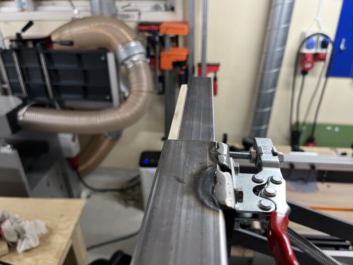 Snabbspännare fäst vid en justerbar metallarm i en verkstad med utsugsrör och verktyg i bakgrunden.