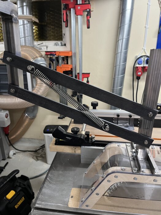 Ett hemmabyggt klingskydd vid en bordssåg, monterat med en arm av metall och fjäderkonstruktion i en verkstadsmiljö.