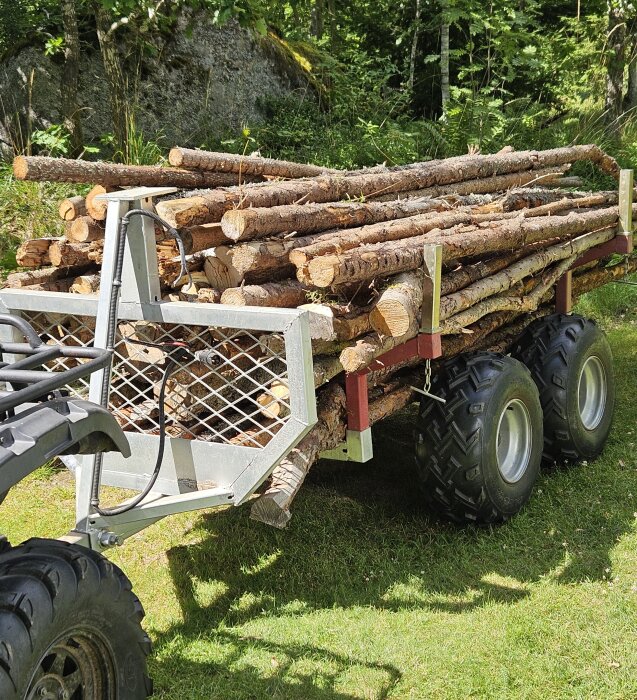 Släpvagn fylld med kaminved, dragen av fyrhjuling, står på en gräsmatta omgivet av skog.