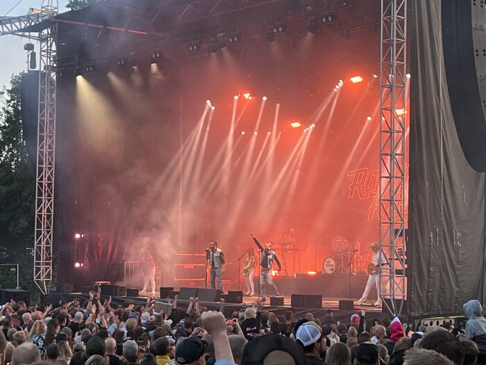 Konsert med band som uppträder på scen inför stor publik, omgiven av färgglatt scenljus och rök, utomhus.